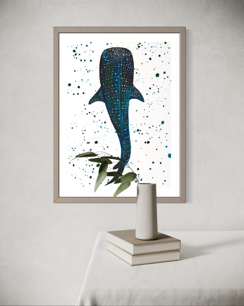 May include: Framed art print featuring a blue whale shark illustration with white spots and a dark blue body. The artwork has a white background with teal paint splatters and green leaves. The frame is a light brown color.