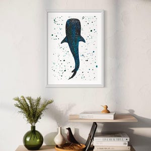 May include: A framed art print featuring a stylized whale shark illustration. The artwork showcases a dark blue whale shark with white spots against a white background speckled with blue and green paint splatters. The print is displayed on a white wall.