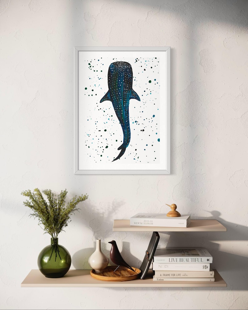 May include: A framed art print featuring a stylized whale shark illustration. The artwork showcases a dark blue whale shark with white spots against a white background speckled with blue and green paint splatters. The print is displayed on a white wall.