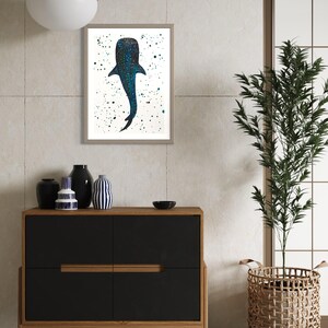 May include: Framed art print featuring a whale shark illustration. The artwork depicts the whale shark in shades of blue with a speckled pattern, set against a white background with scattered green and blue paint splatters. The frame is a neutral color.
