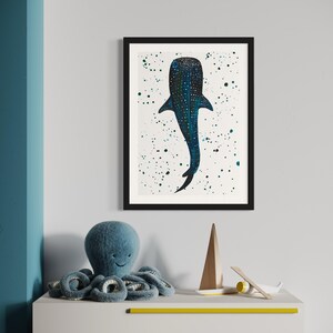 May include: A framed art print featuring a blue whale illustration with white speckles on a white background. The artwork is in a black frame. A blue plush octopus toy, a wooden sailboat, and a small airplane are on a white dresser with yellow accents.