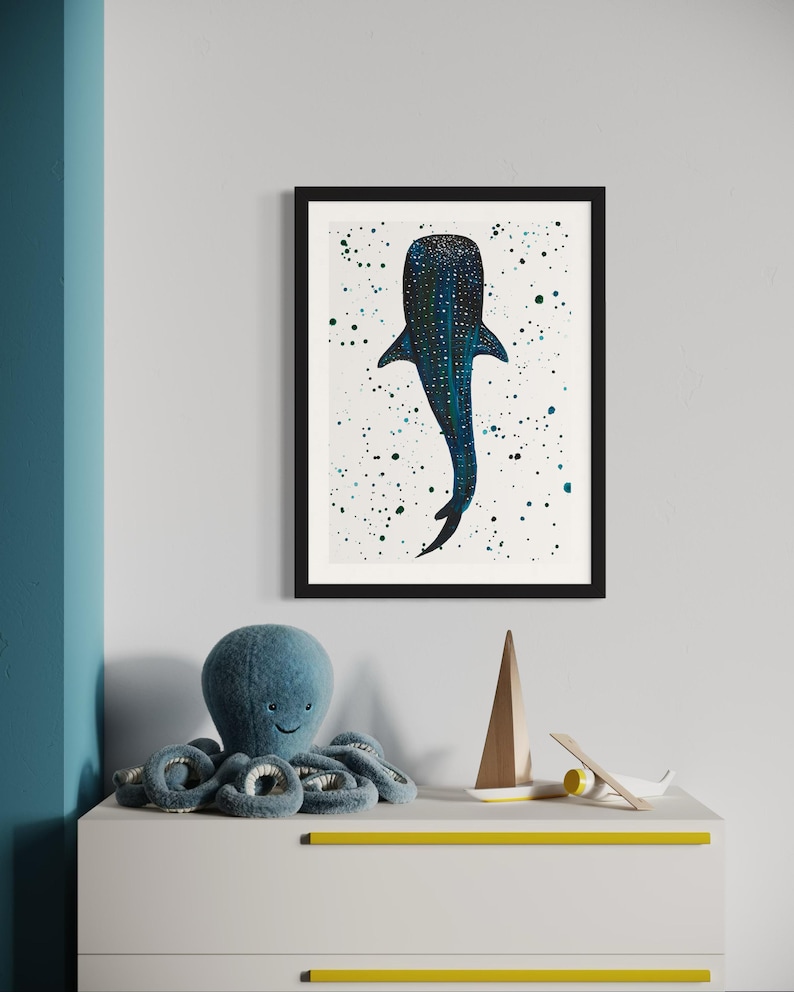 May include: A framed art print featuring a blue whale illustration with white speckles on a white background. The artwork is in a black frame. A blue plush octopus toy, a wooden sailboat, and a small airplane are on a white dresser with yellow accents.