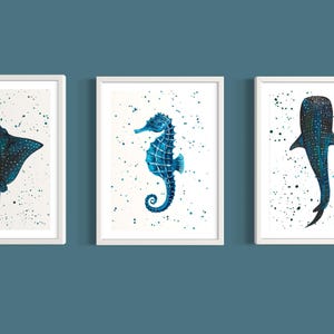 May include: Three framed art prints featuring watercolor illustrations of marine life. The set includes a stingray, a seahorse, and a whale shark, each in shades of blue and teal against a white background with speckled accents.