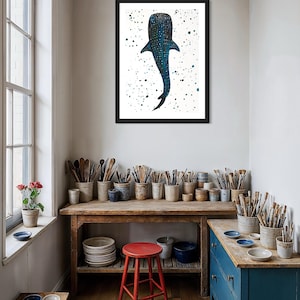 May include: A framed art print featuring a blue whale shark against a white background speckled with dark blue and green dots. The artwork is displayed in a room with a wooden table, red stool, and various art supplies.