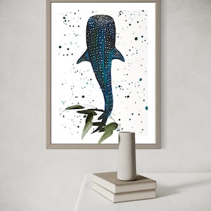 May include: Framed art print featuring a blue whale shark illustration with white spots and a textured pattern. The artwork includes green foliage and a white background with blue and green paint splatters. The frame is a neutral color.