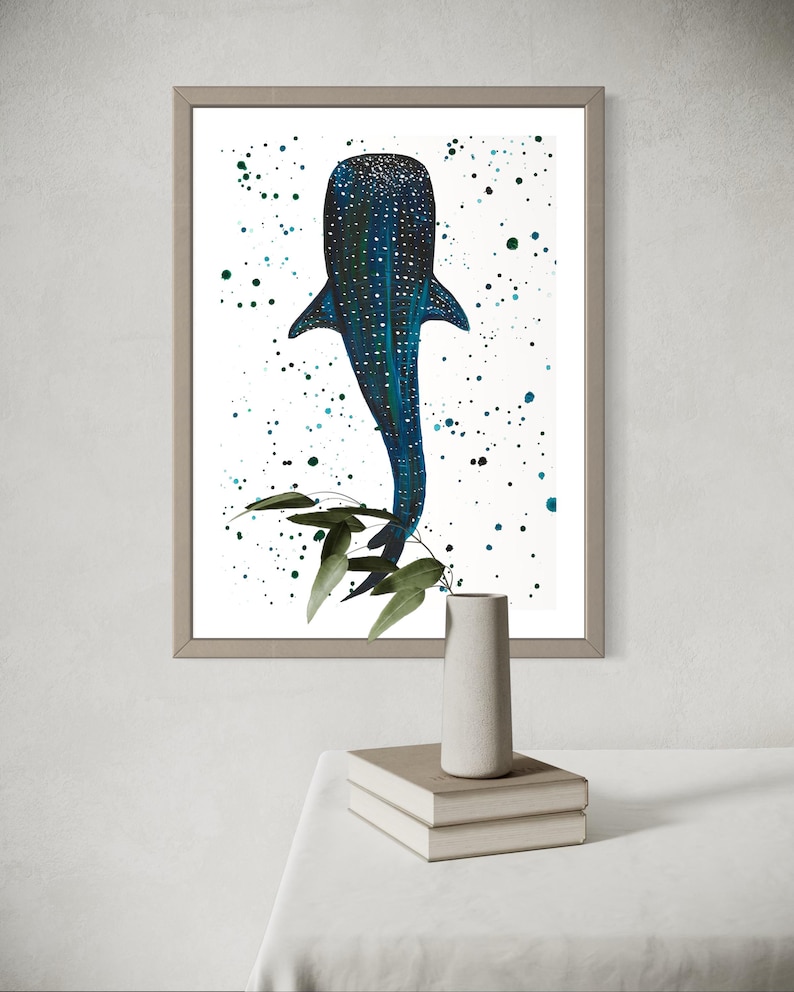 May include: Framed art print featuring a blue whale shark illustration with white spots and a textured pattern. The artwork includes green foliage and a white background with blue and green paint splatters. The frame is a neutral color.