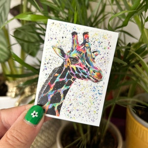 May include: A colourful giraffe sticker with a vibrant, rainbow-hued head and neck, set against a white background speckled with paint. The giraffe's body features a black and white pattern. The sticker is held in front of a green plant.