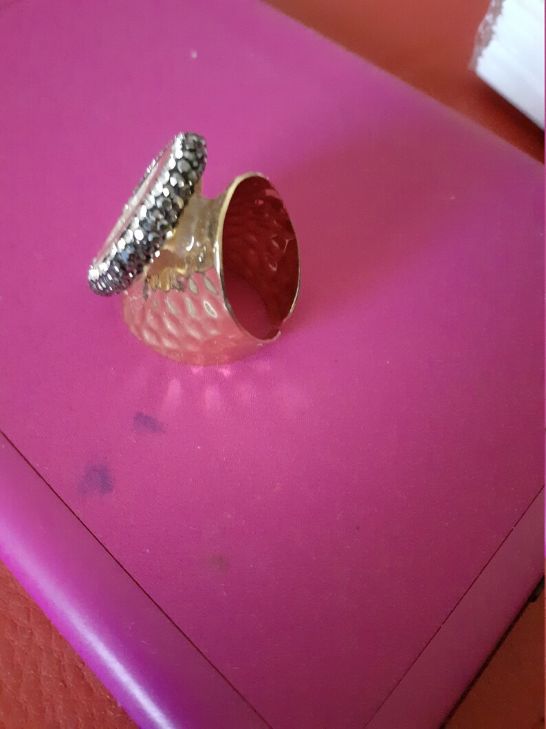 Op de afbeelding: Een gouden ring met een getextureerde band en een zwarte stenen zetting. De ring ligt op een roze oppervlak.