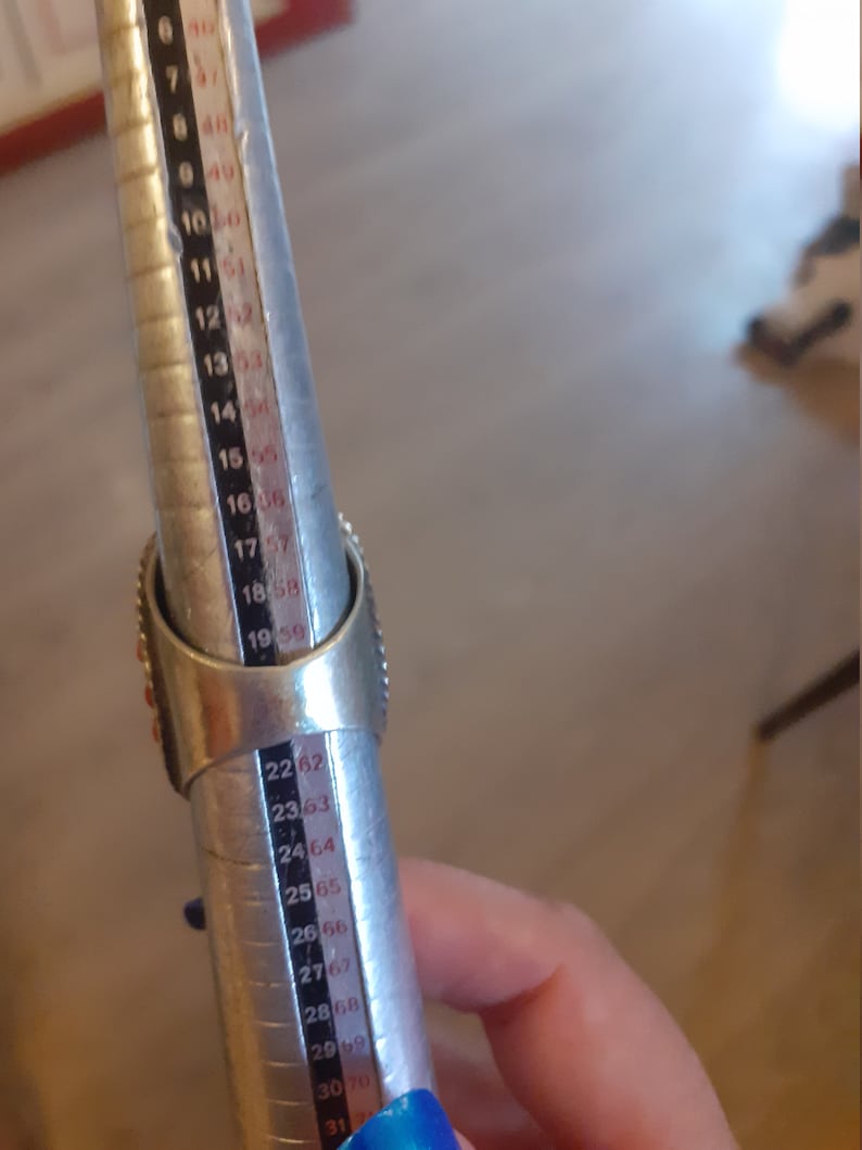May include: A silver ring sizer with red and white stones. The sizer is marked with numbers from 6 to 31.