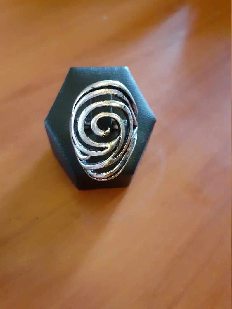 Może przedstawiać: Srebrny pierścionek z wzorem spiralnym. Pierścionek jest prezentowany na czarnym sześciokątnym stojaku.