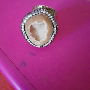 Op de afbeelding: Een zilveren ring met een grote ovale steen. De steen is een lichtbruine agaat met een witte kristallen geode erin. De ring is bezet met kleine heldere strasssteentjes rond de rand van de steen.