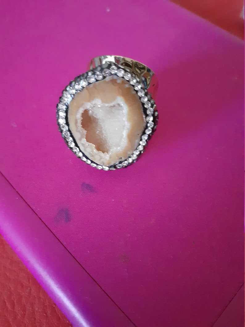 Op de afbeelding: Een zilveren ring met een grote ovale steen. De steen is een lichtbruine agaat met een witte kristallen geode erin. De ring is bezet met kleine heldere strasssteentjes rond de rand van de steen.