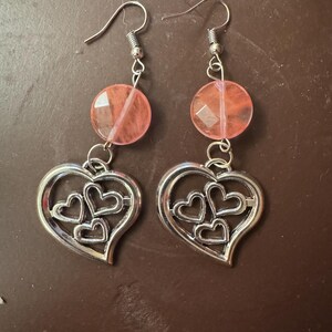 Può includere: Un paio di orecchini pendenti con perline sfaccettate color pesca e ciondoli a forma di cuore argentati. Ogni ciondolo ha un contorno a forma di cuore con tre cuori più piccoli all'interno. Gli orecchini hanno ganci per le orecchie color argento.
