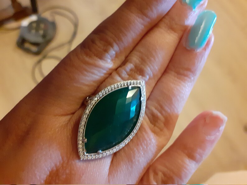 Puede incluir: Un anillo de plata con una gran piedra preciosa facetada de color verde oscuro engastada en un engaste de plata. La piedra preciosa est&aacute; rodeada por una fila de peque&ntilde;as piedras redondas transparentes.