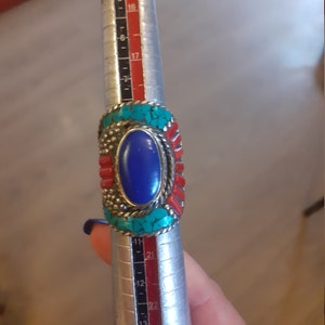 May include: A silver ring with a blue oval stone surrounded by turquoise and red stones. The ring is being sized on a metal ring sizer.