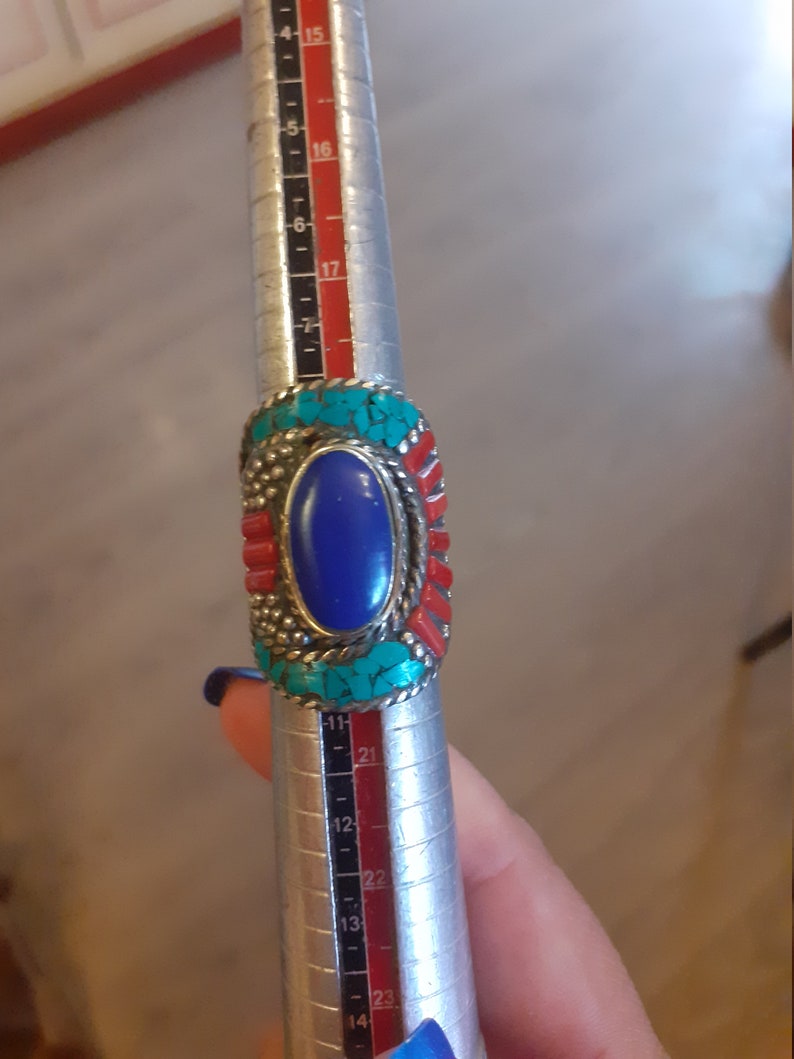 May include: A silver ring with a blue oval stone surrounded by turquoise and red stones. The ring is being sized on a metal ring sizer.
