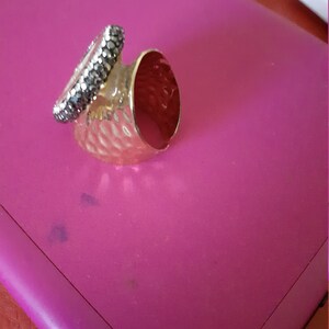 Op de afbeelding: Een gouden ring met een getextureerde band en een zwarte stenen zetting. De ring ligt op een roze oppervlak.