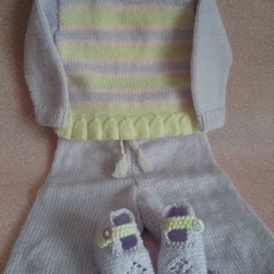 May include: A knitted baby outfit in shades of lavender and yellow. The set includes a striped sweater, pants with ruffles, and matching booties.
