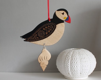 May include: A wooden ornament of a puffin with a black and white patterned body and a red beak. The puffin is hanging from a red string and has a small wooden seashell hanging from its feet.