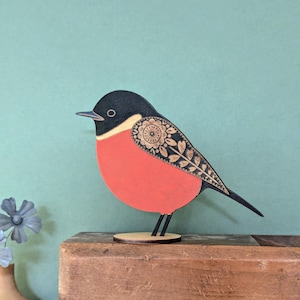 May include: Wooden bird figurine with a black head, a red body, and a decorative floral and leaf design on its wing. The bird stands on a small, round wooden base. The background is a solid teal color.
