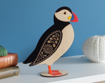 May include: A wooden, black and white patterned puffin figurine with a red and blue beak. The figurine is standing on a wooden base.