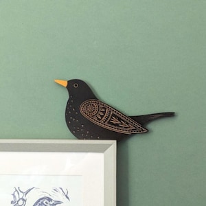 May include: A black wooden bird decoration with an orange beak and detailed etched wing patterns. The bird sits atop a white framed picture against a green wall, offering a nature-inspired decorative element.