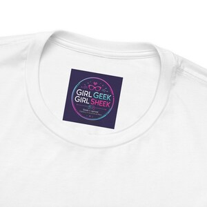 May include: A white t-shirt featuring a dark purple square label on the inside neck area. The label displays a circular logo with pink and blue gradients, pink glasses, a game controller icon, and the text Girl Geek Girl Sheek Geek Inspired.