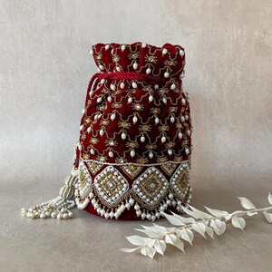 Puede incluir: Una bolsa de cordón de terciopelo burdeos con intrincados bordados de oro y perlas. La bolsa tiene un largo borla de perlas.