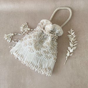 May include: A white beaded drawstring bag with a fringe trim and a pearl-like finish. The bag has a long handle and a tassel.