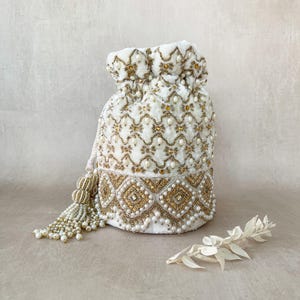 Puede incluir: Una bolsa de cordón de terciopelo blanco con bordado de cuentas doradas y blancas. La bolsa tiene un borla hecha de cuentas blancas.