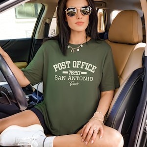 Puede incluir: Camiseta verde oliva con las palabras "POST OFFICE" sobre "78257 SAN ANTONIO Texas" en letras blancas. La persona en el coche lleva gafas de sol y joyas.