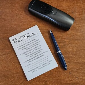 May include: A black telephone, a blue pen, and a white notepad with the text "Customer call: 123 Main St." are on a wooden surface. The notepad has a checklist with various statements.