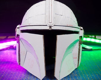 May include: A white cardboard helmet with a black visor, resembling a Mandalorian helmet. The helmet is partially assembled and has visible numbers and markings.