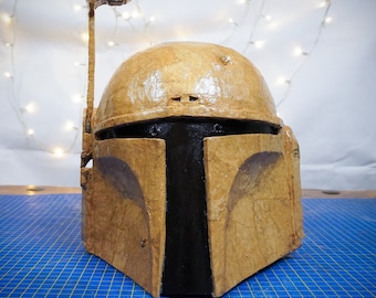 May include: A brown and black Star Wars Boba Fett helmet made of papier-mâché. The helmet has a black visor and a brown, textured surface.
