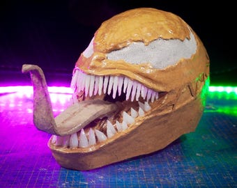 Könnte beinhalten: Eine braune und weiße Papiermaché-Maske von Venom mit einem großen, offenen Mund und einer langen, braunen Zunge, die herausragt. Die Maske befindet sich auf einer blauen Schneidematte mit einem violetten und grünen Licht dahinter.