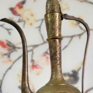 May include: An antique brass pitcher with intricate floral engravings. The pitcher features a long, curved spout and a handle. The surface has a warm, golden hue, and the design suggests an Eastern influence. The pitcher is likely used for decorative purposes.