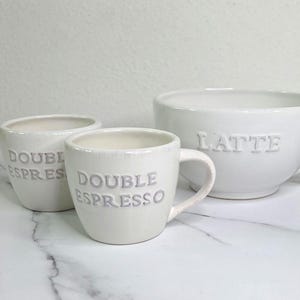 May include: A set of three white ceramic coffee mugs and bowls. Two smaller mugs are labeled "DOUBLE ESPRESSO," and a larger bowl is labeled "LATTE." The mugs and bowl have a simple, elegant design.
