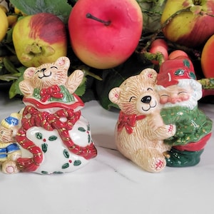 May include: Two ceramic Christmas figurines: a bear in a gift bag and a bear hugging a gnome. The bear in the bag is white with red and gold accents. The hugging bear has a red bow. The gnome wears a green coat.