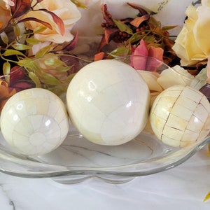 May include: Three white decorative balls with a textured surface are displayed in a clear glass bowl. The bowl has a wavy edge and is sitting on a white surface.