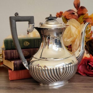 May include: A silver teapot with a black wooden handle and a decorative, fluted design. The teapot is sitting on a stack of books and a wooden surface.