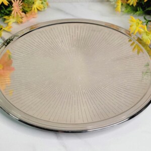 May include: A round, silver-rimmed serving tray with a radial pattern emanating from the center. The tray has a light beige surface and is surrounded by a silver border. Decorative flowers are in the background.