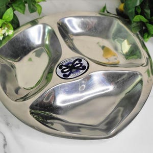May include: A stainless steel divided serving dish with three compartments. The dish has a decorative floral design in the center. The dish is round with a curved shape.