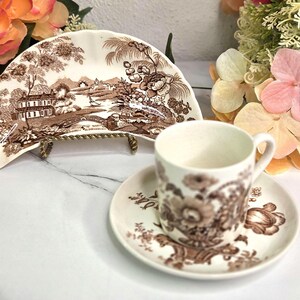 May include: A ceramic demitasse cup and saucer set featuring a brown floral transferware pattern, accompanied by a matching crescent-shaped side plate displayed on a small gold-toned metal stand, all decorated with detailed landscape and floral motifs.
