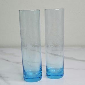 May include: Two tall, clear drinking glasses with a light blue base and a subtle etched design. The glasses have a cylindrical shape and are displayed against a neutral background. These glasses are suitable for serving water or other beverages.