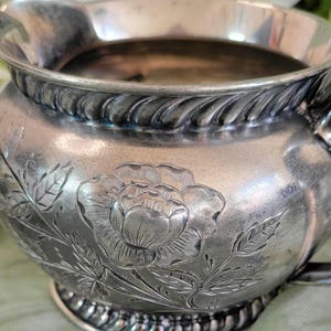 May include: Silver creamer with a floral design featuring a large rose and leaves. The creamer has a rope-like design around the top and bottom.