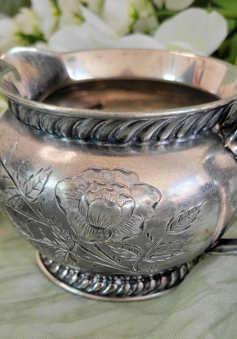 May include: Silver creamer with a floral design featuring a large rose and leaves. The creamer has a rope-like design around the top and bottom.