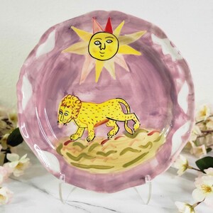 May include: A ceramic plate with a pink background and a yellow lion walking across a green and brown landscape. A yellow sun with a face and a red hat is above the lion.