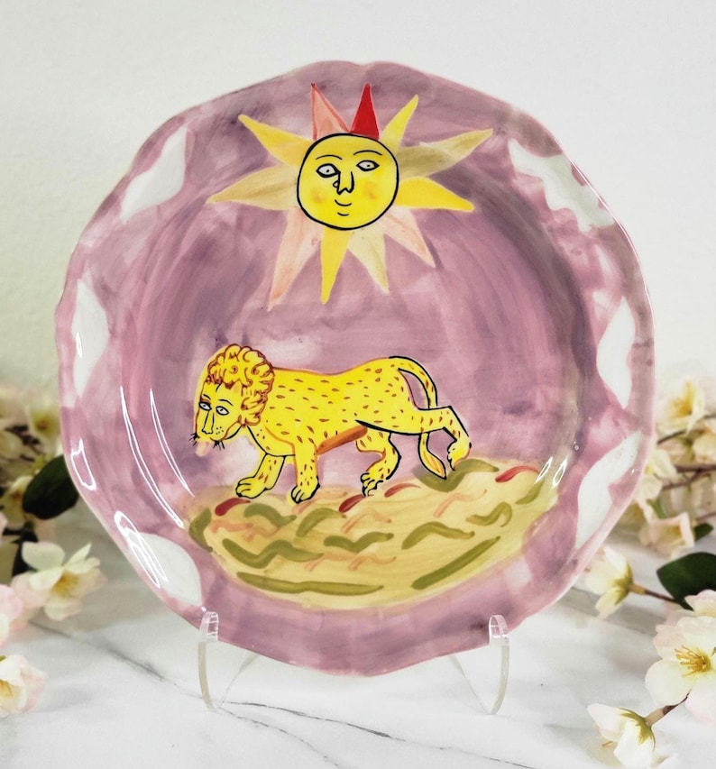 May include: A ceramic plate with a pink background and a yellow lion walking across a green and brown landscape. A yellow sun with a face and a red hat is above the lion.
