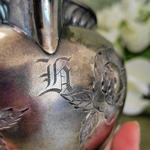May include: Silver creamer with an engraved initial "H" and floral design. The creamer has a rope-like design around the base.