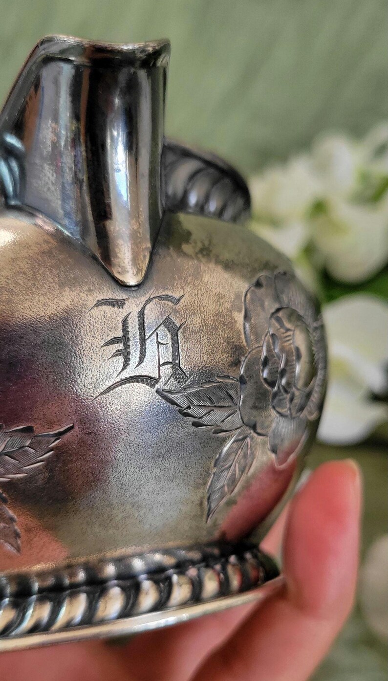 May include: Silver creamer with an engraved initial "H" and floral design. The creamer has a rope-like design around the base.